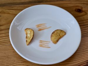Two fried potato wedges arranged to resemble eyes and a mouth on a white plate, creating a fun, smiley face. A few lines of sauce add expression. Taken at a café near Fort Kochi Beach on a wooden table.  