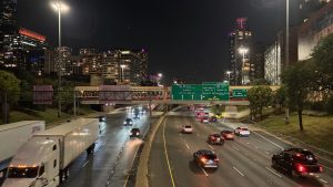 2 5-lane freeways, seen from a bridge over them. There are large green signs over the roadway. There are sky scrapers in the distance.  This is a night scene in Chicago.