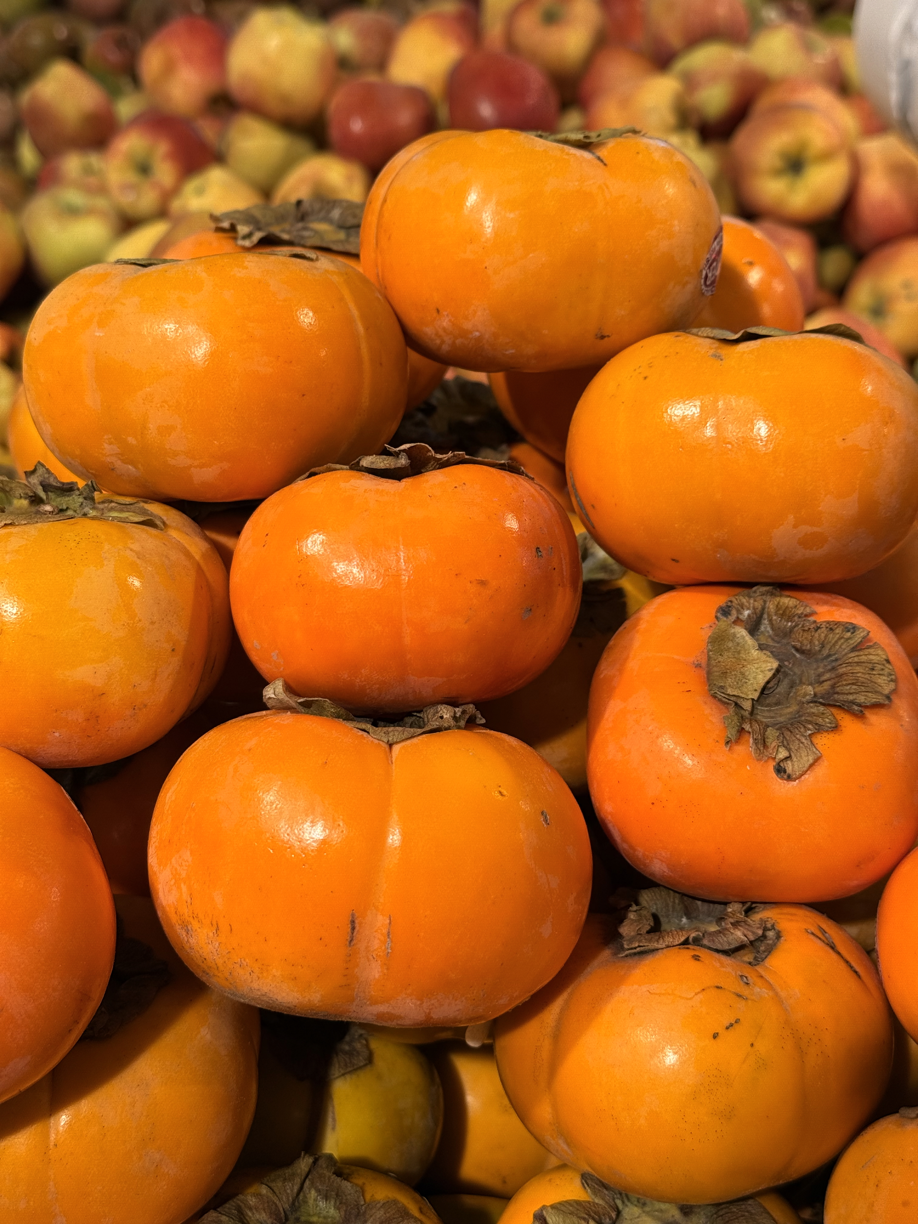 A fresh pile of orange persimmons is stacked in a market fruit display. Red and green apples in the background. Captured at a vegetable shop in Palazhi, Kozhikode.