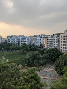 View of a park and tall residential buildings of similar heights, showcasing greenery of trees and green lake in the middle of apartments.