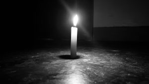 A single candle is burning in a dark setting, casting a soft glow on the surface beneath it with a flame illuminating the area around it while the background fades into shadow, creating a moody and atmospheric scene.