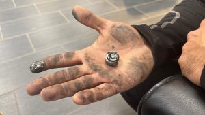 A hand, mostly filling the screen held open palm up.  The hand is quite dirty. In the center of the palm is a metal plug with grease on it, and on the index finger is a silver paste.