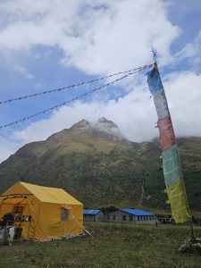 A vibrant yellow tent stands in the foreground, with a person partially visible inside. Behind the tent, there are stone buildings with blue roofs, set against a backdrop of steep mountains shrouded in clouds. Colorful prayer flags flutter in the breeze near the tent, adding a cultural touch to the serene mountain landscape.