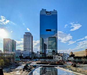 A modern glass skyscraper towers in the background, reflecting the blue sky and nearby buildings. In the foreground, there are construction sites with machinery and scattered materials.