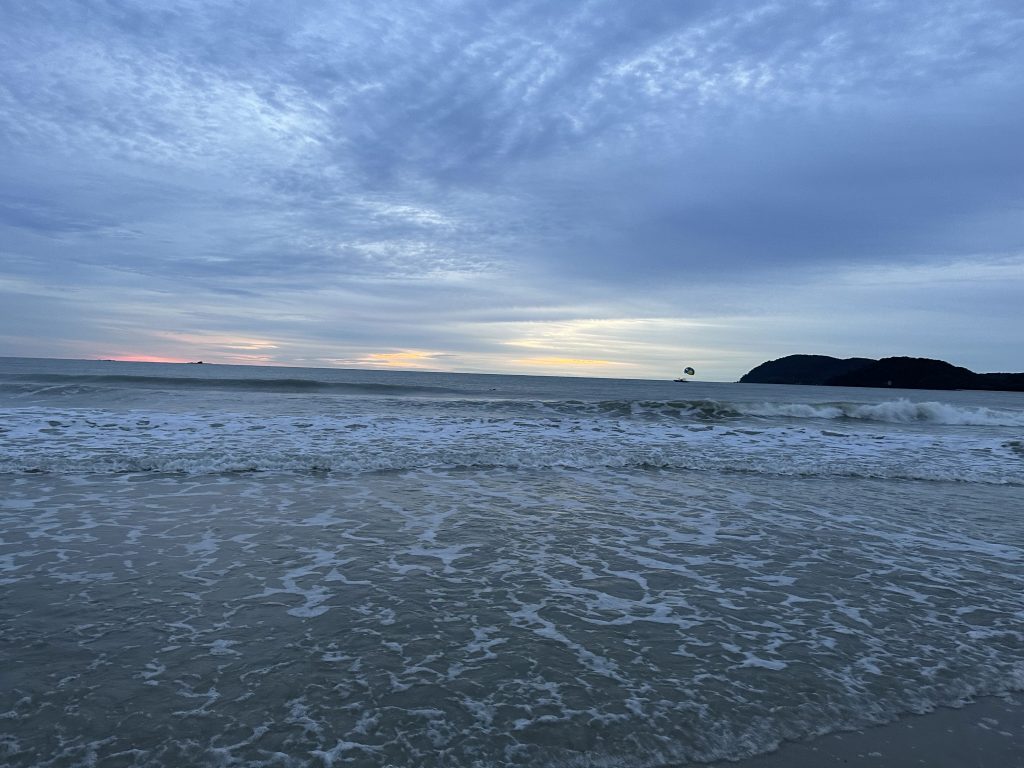 A peaceful evening view of the beach in Langkawi, Malaysia, with gentle waves washing onto the shore and soft sunset colors on the horizon. Distant islands and a parasail in the sky add depth to this serene coastal scene.