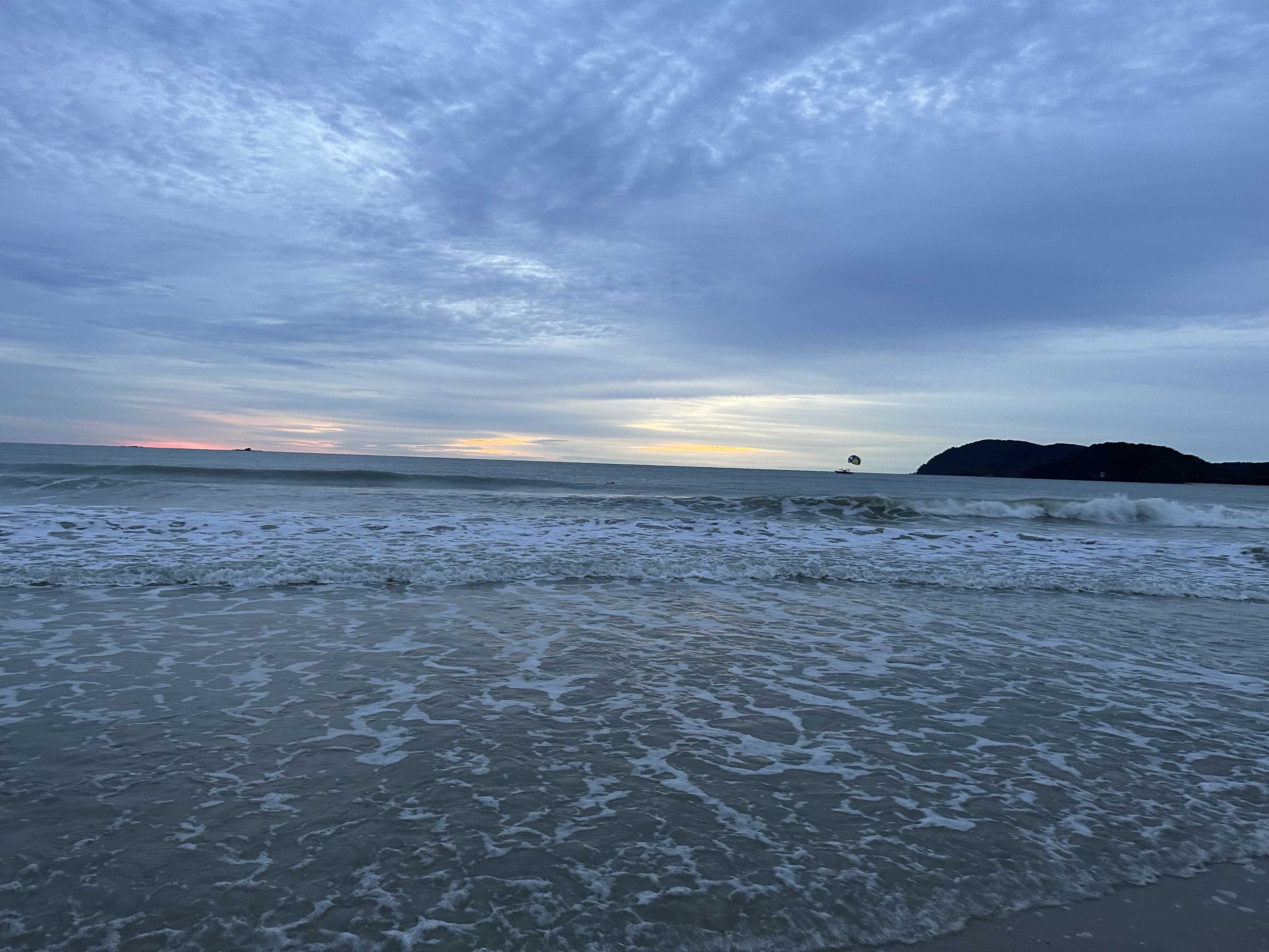 A peaceful evening view of the beach in Langkawi, Malaysia, with gentle waves washing onto the shore and soft sunset colors on the horizon. Distant islands and a parasail in the sky add depth to this serene coastal scene.