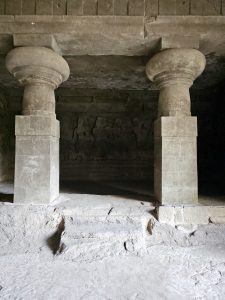 An interior view of a cave, featuring thick stone pillars and faint wall carvings. Shot at Elephanta Caves, Mumbai, Maharashtra.
