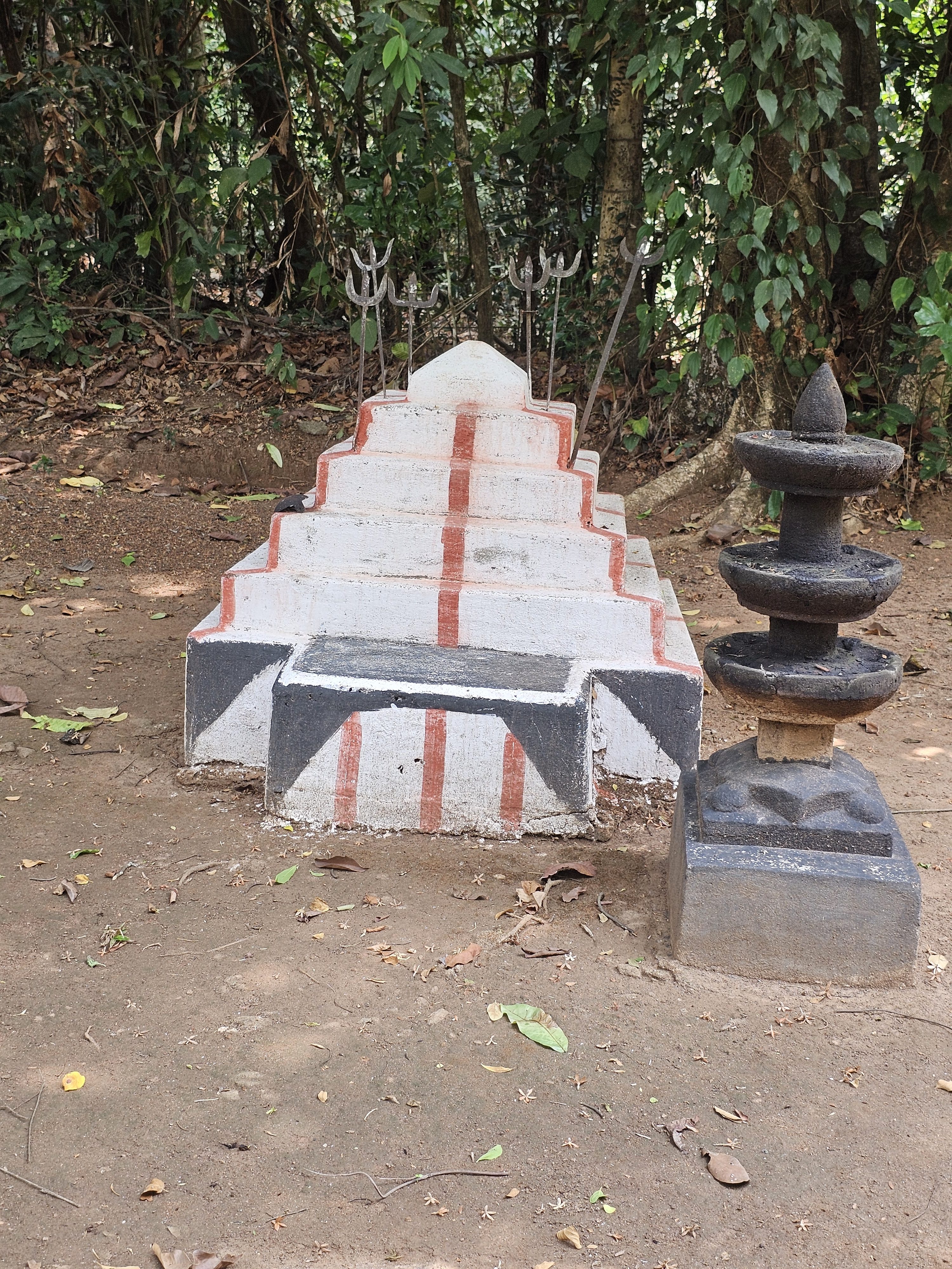 A small white and red shrine with tridents placed behind it, located in a forest area. The structure stands on bare ground. Captured from Wandoor, Kerala.