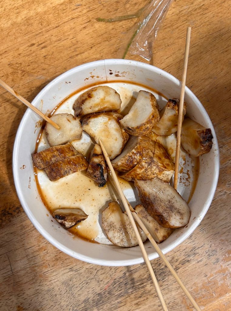 A white bowl containing several pieces of grilled or sautéed fish or meat, coated in a dark sauce, with some pieces on wooden skewers.
