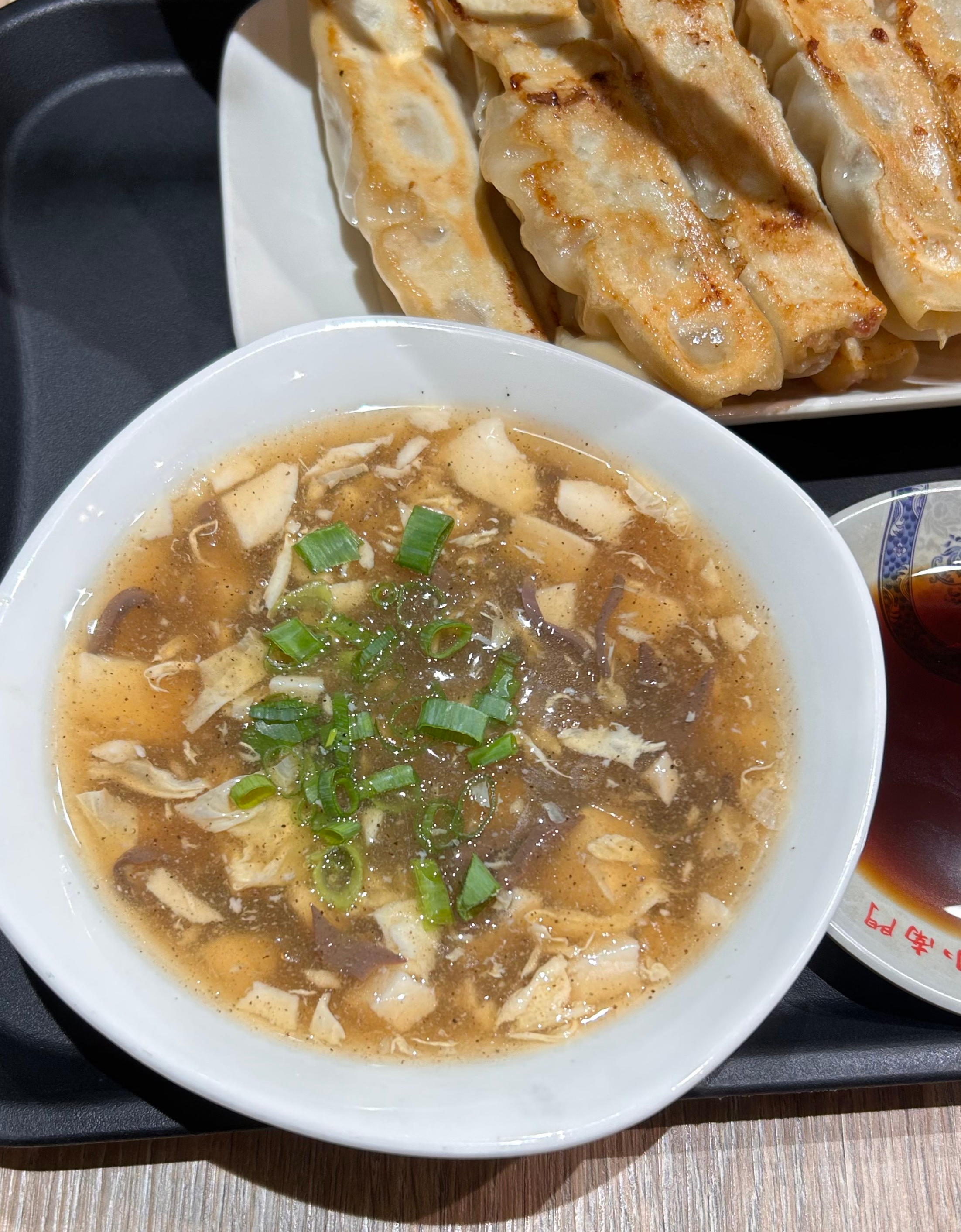 A bowl of savory broth with chopped tofu, shredded meat, and green onions is placed on a black tray.