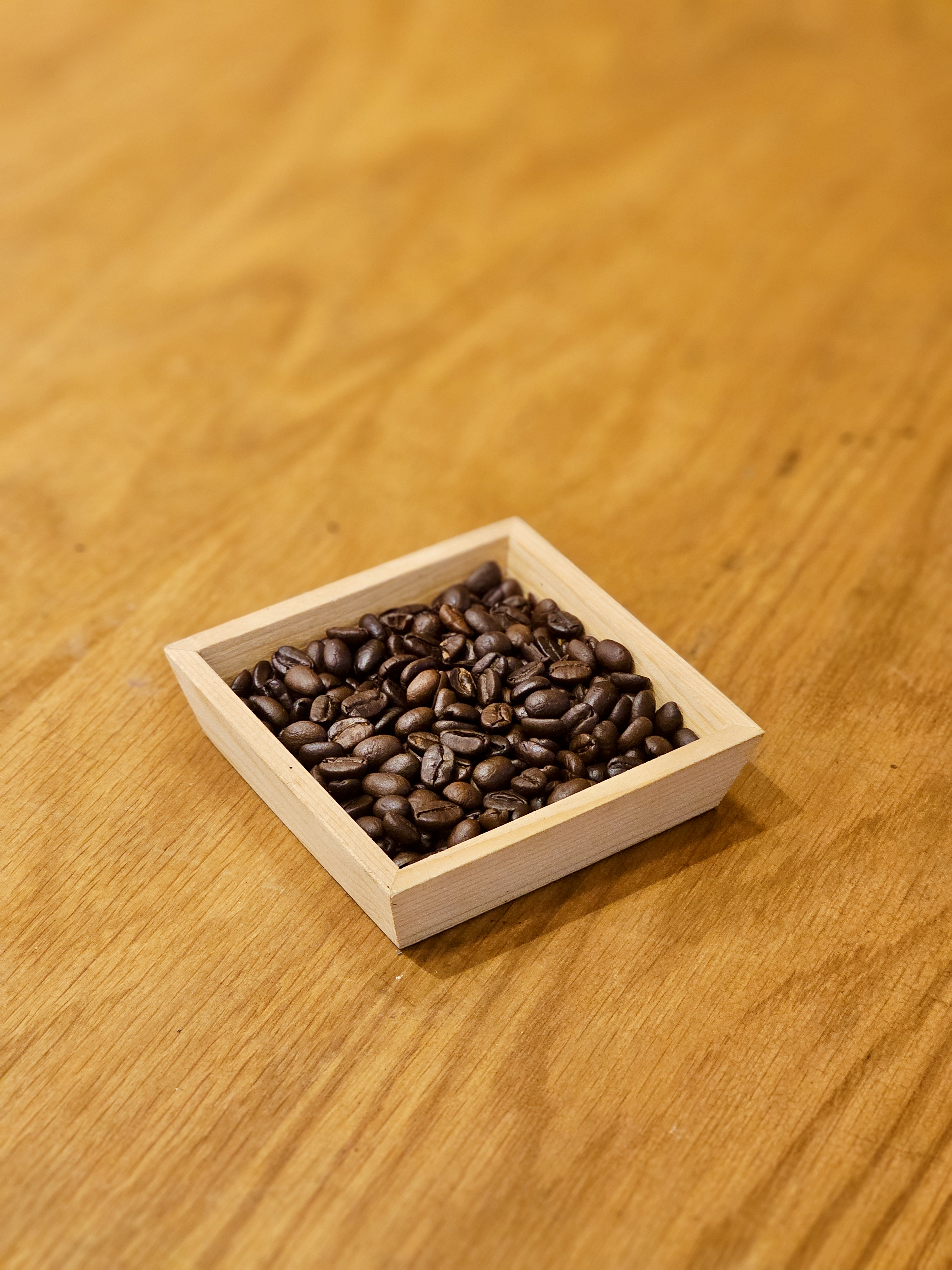 A small wooden tray filled with dark roasted coffee beans sits on a smooth wooden table in Kozhikode, Kerala. The warm tones make the beans look rich and fresh.
