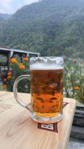 A close-up view of a frosty glass mug filled with amber beer, resting on a wooden surface with the blurry background of  green hills and flowers.