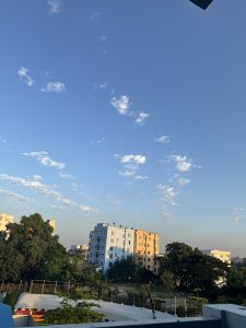 A clear blue sky dotted with scattered white clouds, overlooking a residential area with several multi-story buildings, some painted in pastel colors. 