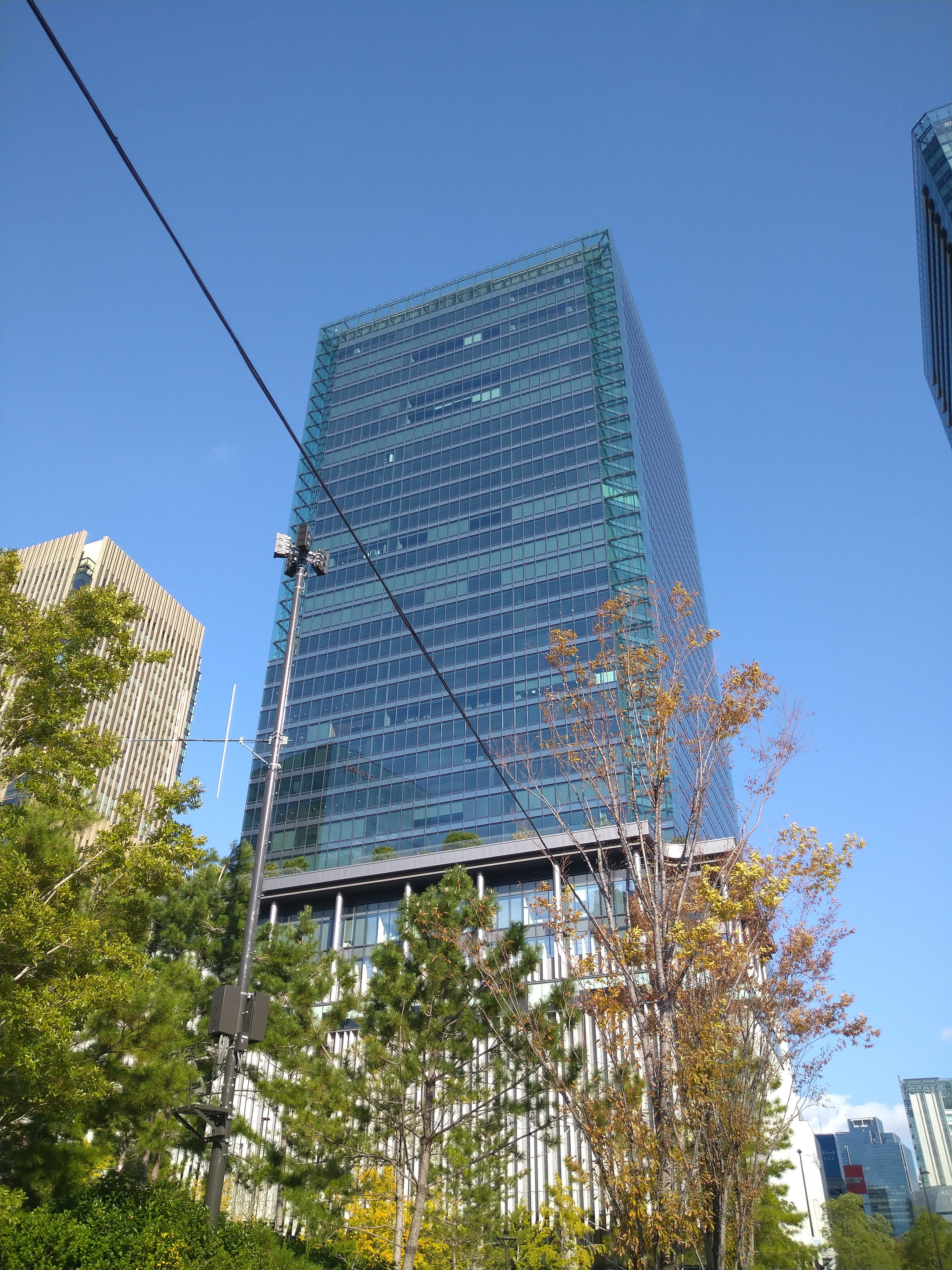 An image of a tall building in osaka with another a bit smaller buildings on its either sides. Its front view has some green trees and top view has a clear blue sky.