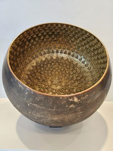 A close-up of a large metal bowl decorated with many tiny golden Buddha figures inside. The bowl is displayed on a white platform, taken from the BKC Complex, Mumbai, Maharashtra. 