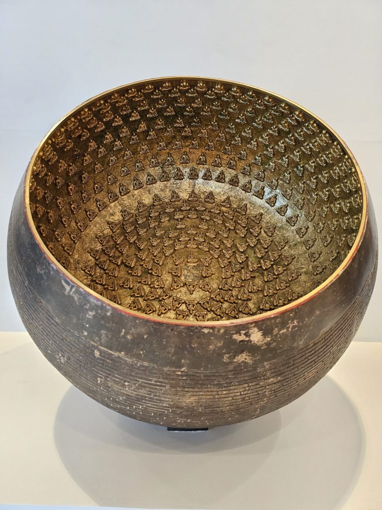 A close-up of a large metal bowl decorated with many tiny golden Buddha figures inside. The bowl is displayed on a white platform, taken from the BKC Complex, Mumbai, Maharashtra.