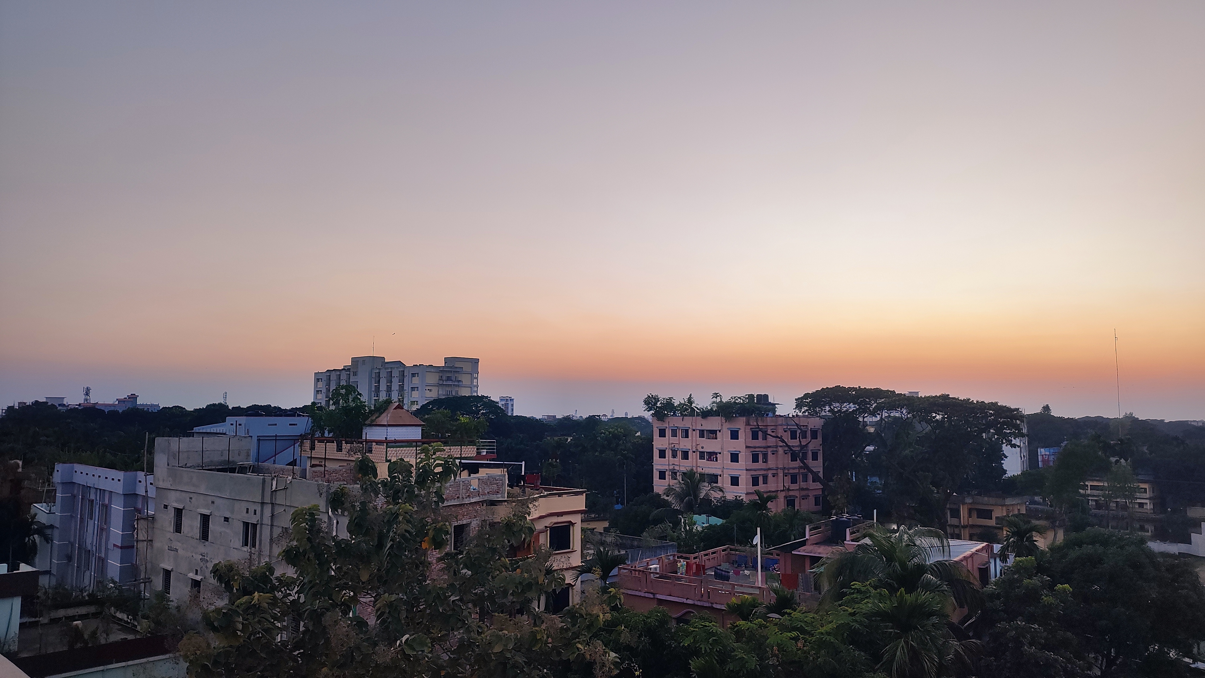 An elevated, wide-angle photo of a cityscape at sunset or sunrise. The sky displays a soft gradient of orange, pink, and pale yellow merging into a light purple-grey. Below, a mixture of low- to mid-rise buildings are visible, interspersed with dense green trees and foliage.