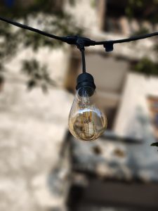 A close-up view of a vintage-style light bulb hanging from a black cord, with a blurred background that includes a partially visible wall and greenery. T