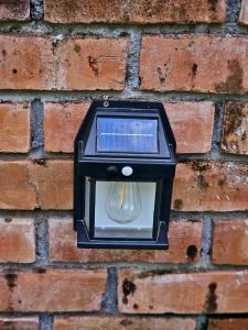 A small solar-powered wall light is fixed on a red brick wall. The black frame and glass bulb stand out clearly. Taken in Perumanna, Kozhikode, Kerala. 