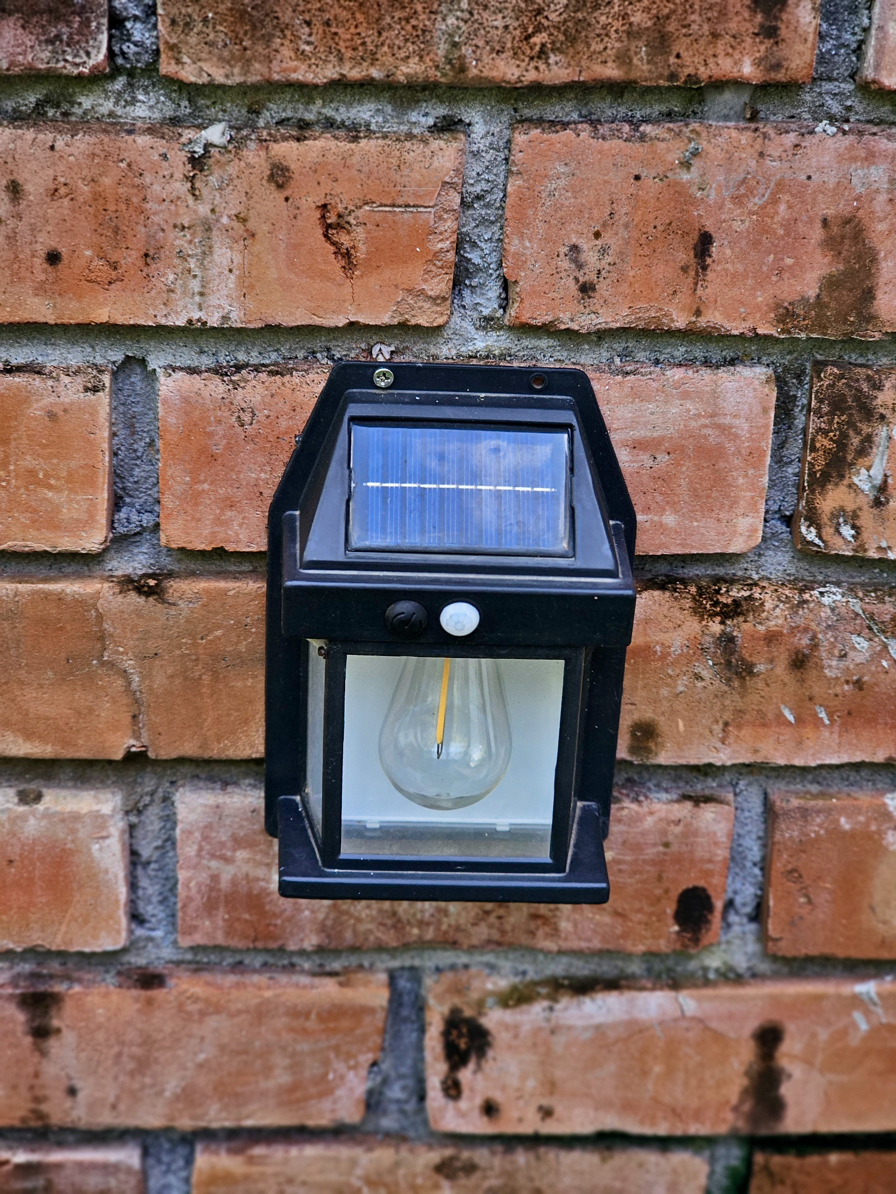 A small solar-powered wall light is fixed on a red brick wall. The black frame and glass bulb stand out clearly. Taken in Perumanna, Kozhikode, Kerala.