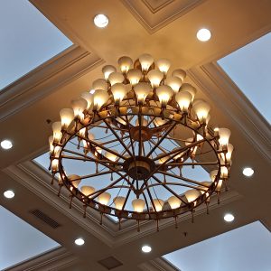 A large, circular chandelier hangs from a ceiling, featuring multiple light bulbs arranged in a radial pattern. The chandelier is made of a golden-brown metal with a vintage design, and it emits a warm light. Surrounding the chandelier are recessed ceiling lights, and above it is a sky blue ceiling section.