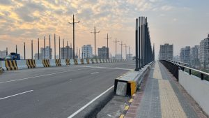 A wide paved bridge with tall black poles leads towards the city skyline under a soft morning sky.

