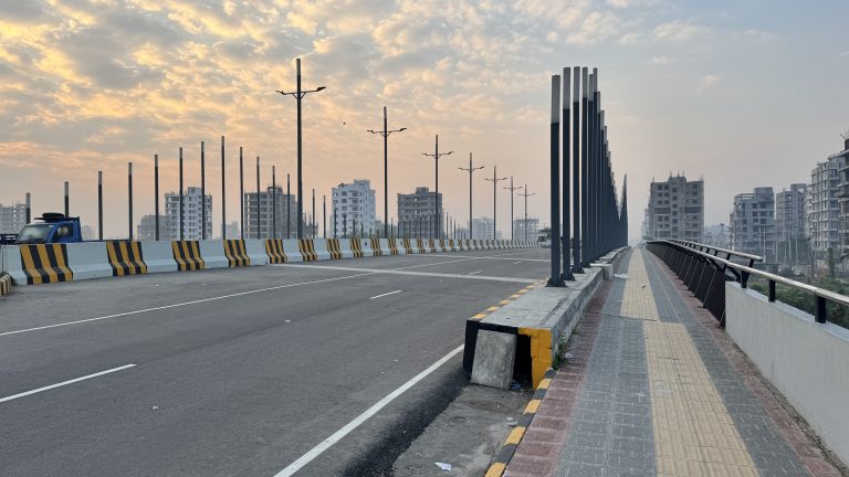 A wide paved bridge with tall black poles leads towards the city skyline under a soft morning sky.