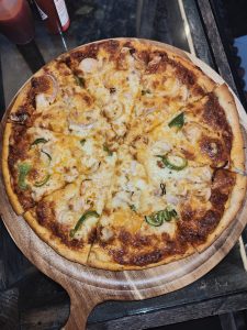 A freshly baked pizza on a wooden serving platter, topped with melted cheese, pieces of chicken, and green bell pepper slices. 