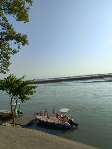 A peaceful riverside scene with two boats at a floating platform—one decorated with many red Nepali flags, the other partly covered by a green tarp