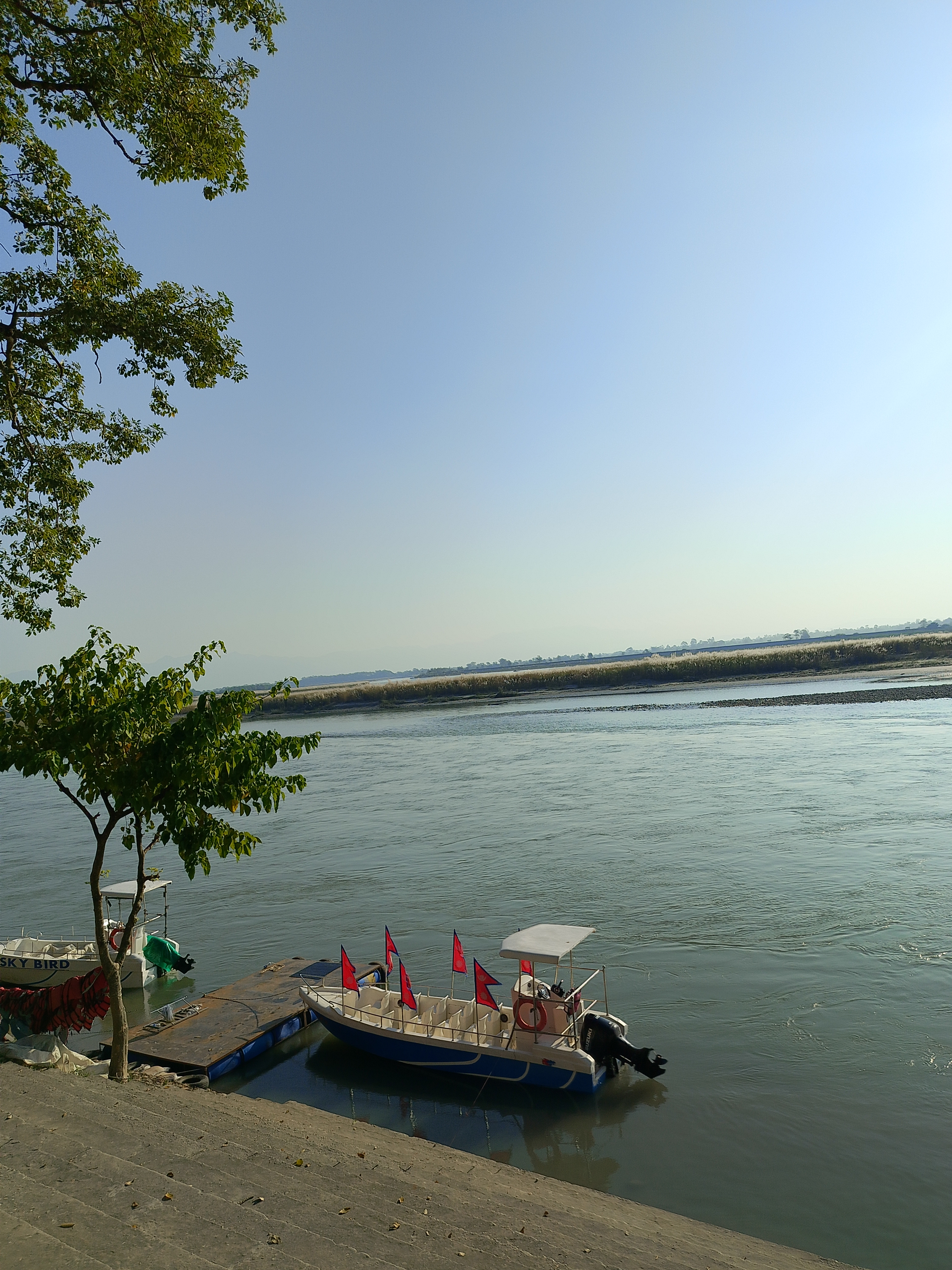 A peaceful riverside scene with two boats at a floating platform—one decorated with many red Nepali flags, the other partly covered by a green tarp