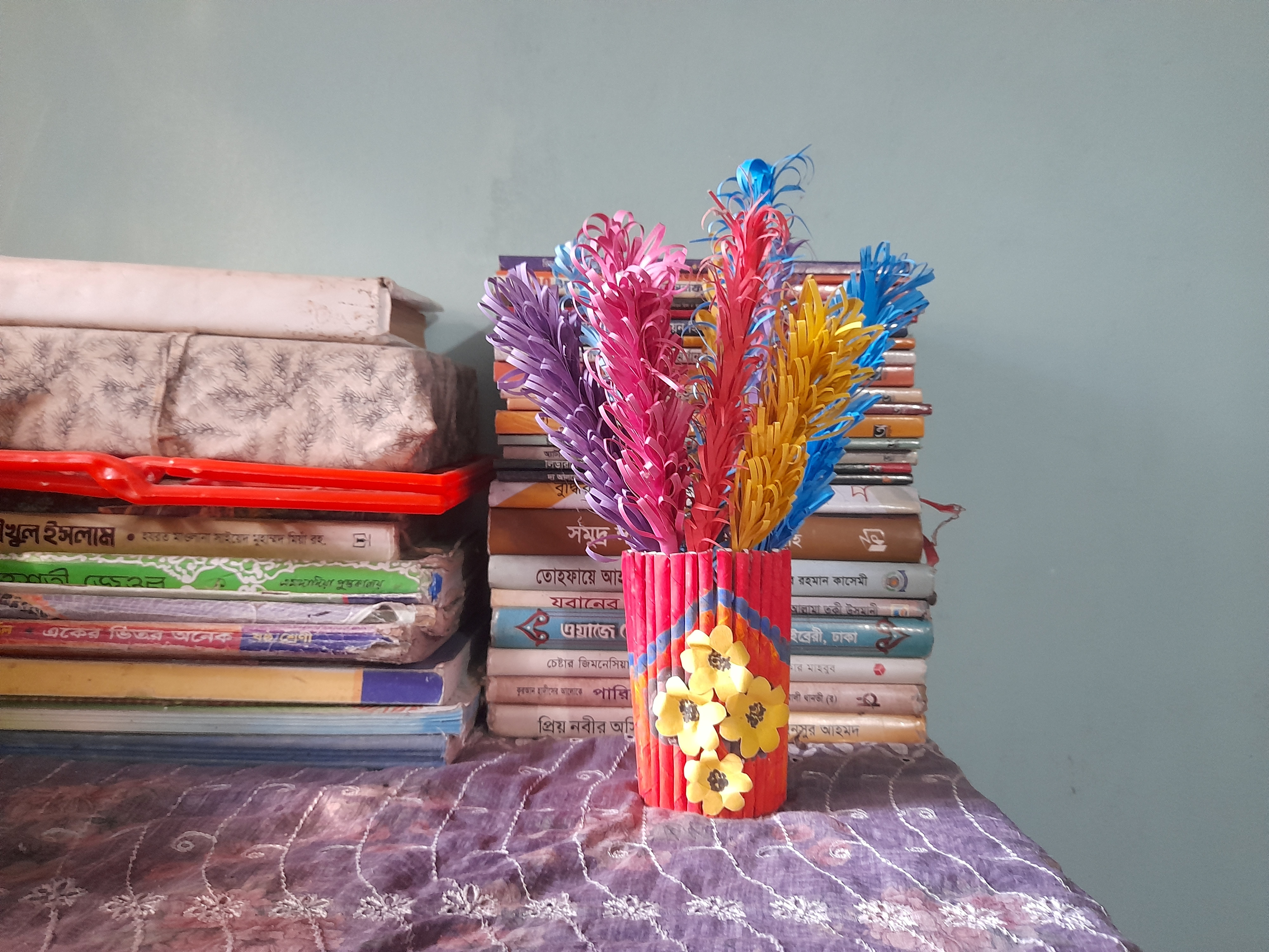Colorful paper flowers in a red, decorated vase are placed on a patterned tablecloth. Behind them is a stack of books against a muted wall. This photo was taken in Harbaid, Gazipur.