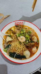 A close-up view of a bowl of spicy noodle soup featuring yellow noodles topped with shrimp, pieces of meat, green onions, and fresh herbs. 
