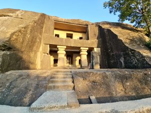 The main entrance of a Kanheri Cave, featuring stone steps, carved pillars, and a large rock face illuminated by warm sunlight, is a beautiful example of ancient Buddhist architecture. 