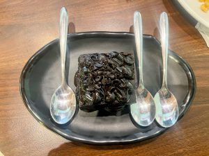 A small, square dark chocolate brownie topped with chocolate chips and glaze is centered on a black, oval platter, flanked by three silver spoons.