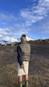 A person stands on a grassy patch, facing away from the camera and facing towards a view of clouds on green hills and some pet goats, wearing a puffy gray jacket, green boots and a white skirt. 