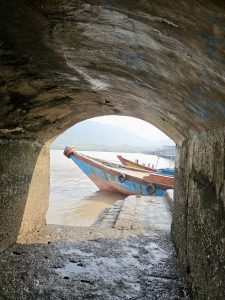 An old wooden boat is seen through a stone arch at the water’s edge. The mountain and shore appear softly in the background. Captured near Elephanta Caves, Mumbai, Maharashtra. 
