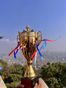 A shiny gold trophy with colorful ribbons in red and blue is being held in front of a scenic view of a city.