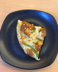 A single slice of pizza is presented on a black plate placed on a brown wooden table.