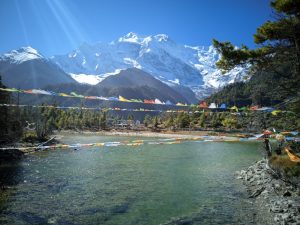 

A peaceful lake rests in a green valley, with snowy peaks and colorful prayer flags fluttering under a bright blue sky