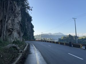 A winding road runs alongside a rocky cliff covered with greenery. 