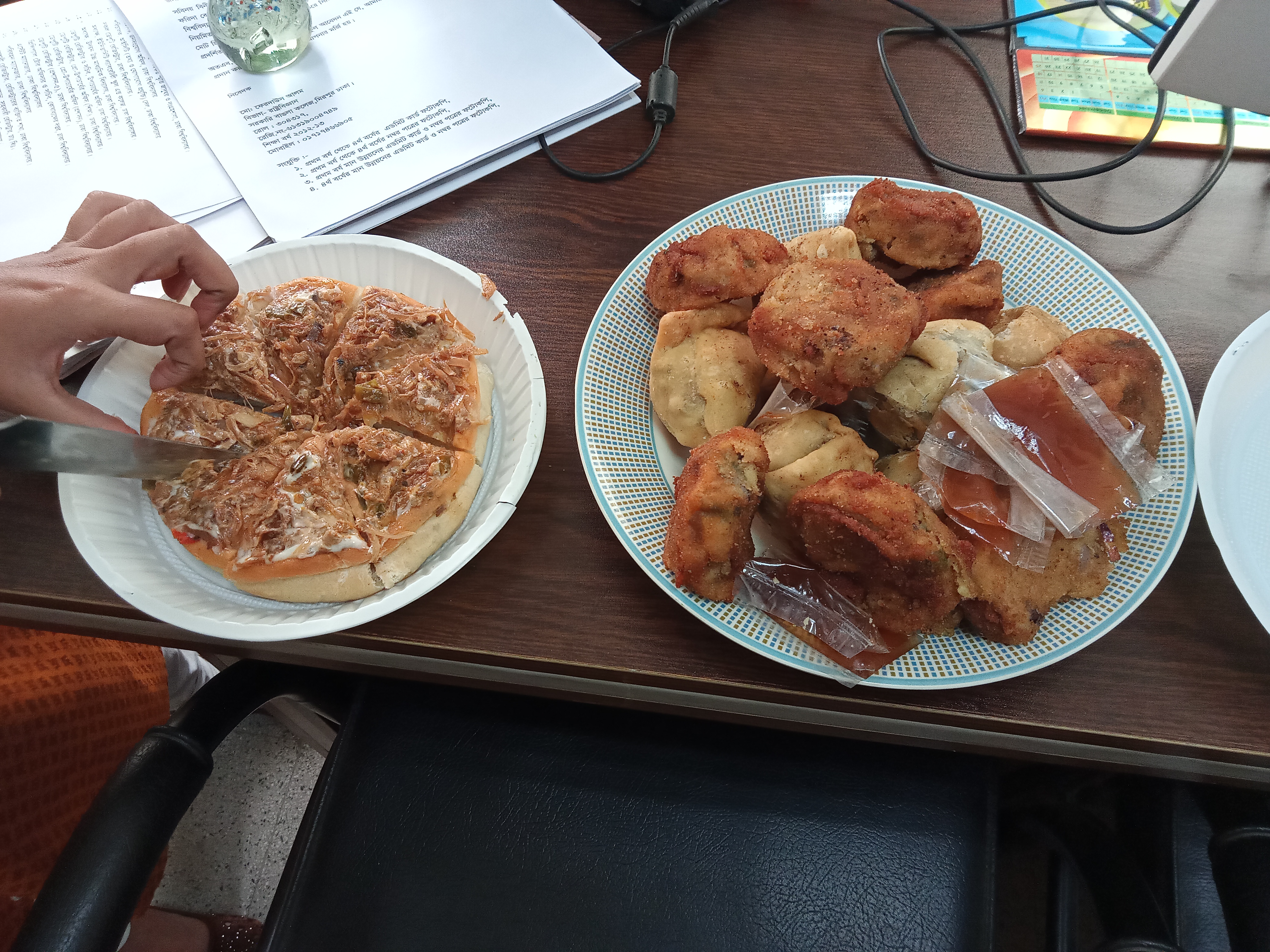 The pizza slice grabbed by a human hand, and the snacks placed on a brown desk. Dhaka, Bangladesh