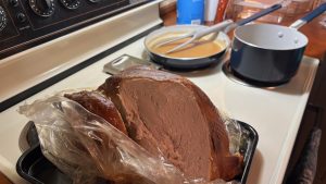 A meal on the stovetop.  In the foreground, a turkey in a plastic bag on a metal plate with a slide taken from the breast. In the background a flat pan with gravy in it, and a saucepan.

