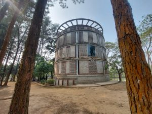 A unique, circular, three-tiered modern building made of gray brick and concrete, framed by tall pine trees on a sunny day at Zinda Park in Bangladesh.
