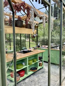 A green kitchen inside a glass house. Copper pots, garlic, and dried peppers hang from wooden beams, and colorful cookware sits on open shelves under a wooden counter.