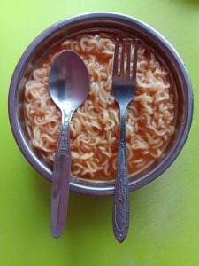 A round metal bowl of instant noodles in reddish sauce with a spoon and fork. Dhaka, Bangladesh.