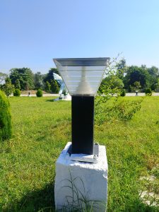 A modern outdoor lamp post stands on a white concrete base, surrounded by green grass and small bushes. 