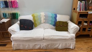A white sofa against a wall in a yarn shop.  There are crocheted pillows and a blanket on the sofa, and skeins of yarn hanging on hooks around it.