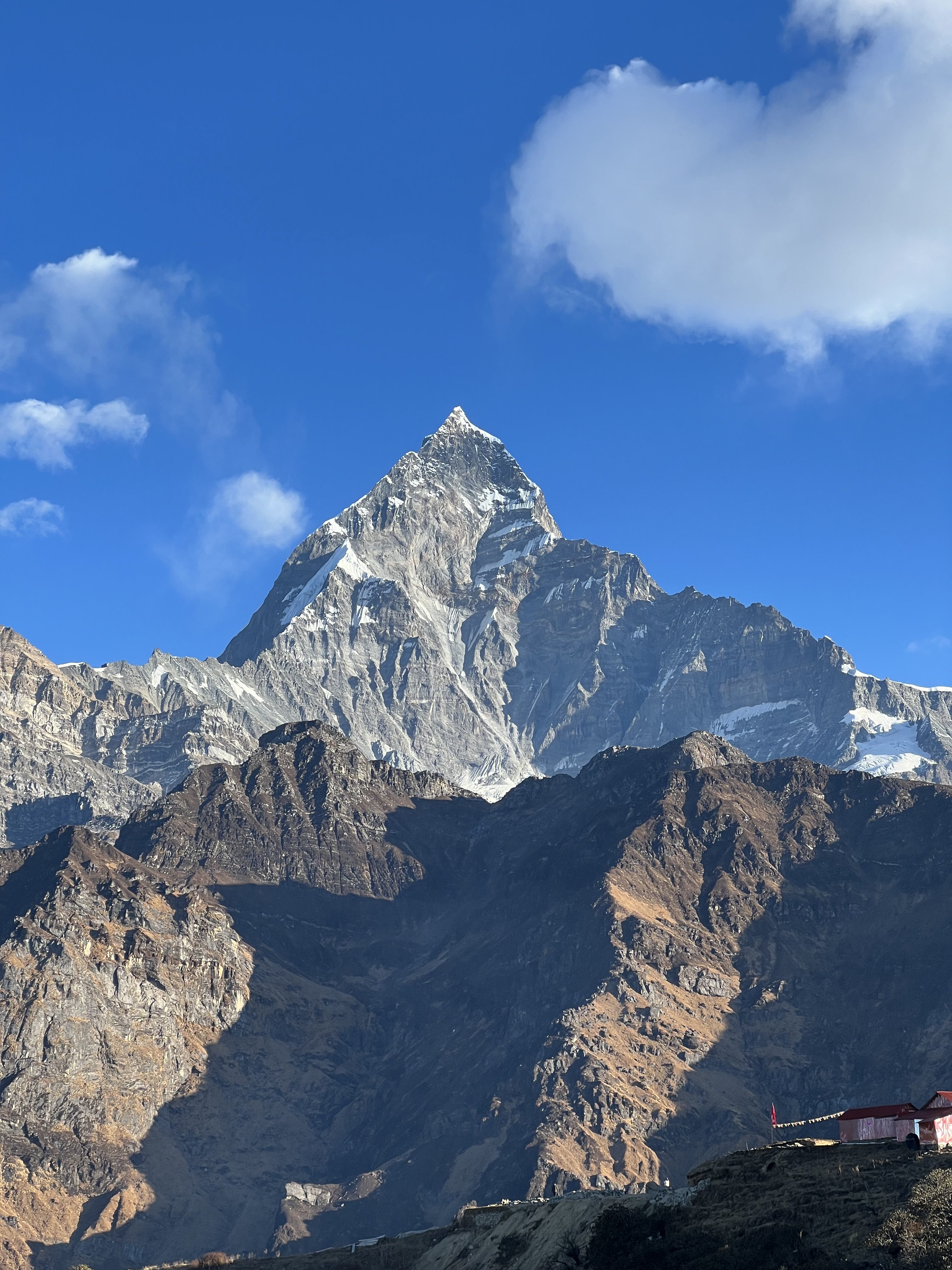 

A majestic Machhapuchhere mountain peak rises sharply against a clear blue sky, with scattered white clouds partially visible. 