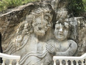 A large stone relief sculpture carved into a rock face depicts a serene woman and a young child. The woman, with long flowing hair, gazes tenderly at the child she holds close. Both figures are sculpted in light-colored stone, weathered with age and streaks of natural patina. A white balustrade runs along the bottom edge of the image, partially obscuring the lower portion of the carving. Lush greenery grows atop and around the rocky surface in the background.