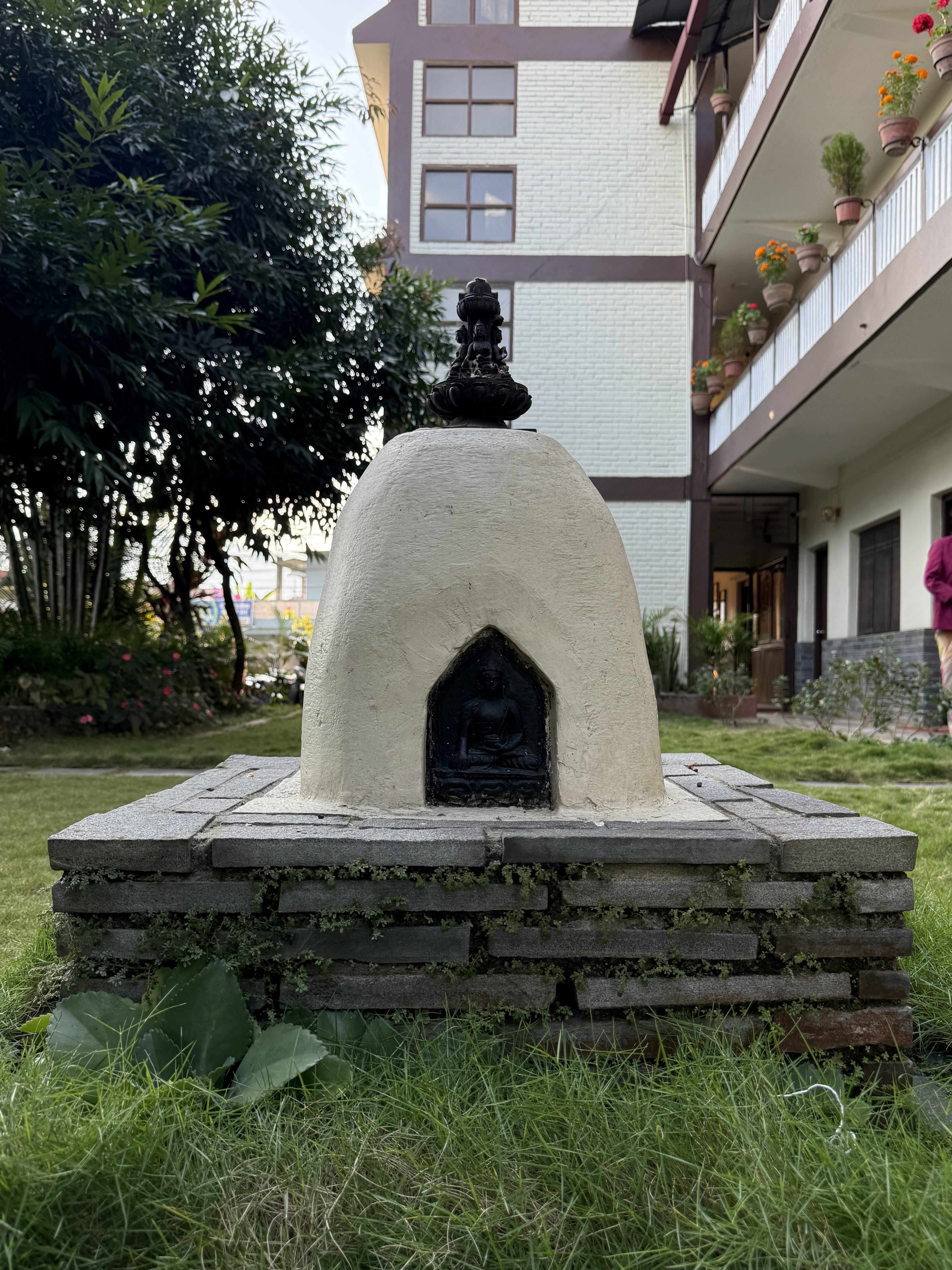 A small white structure with a dome shape sits on a stone base, featuring a black entrance where a statue of Buddha is visible.
